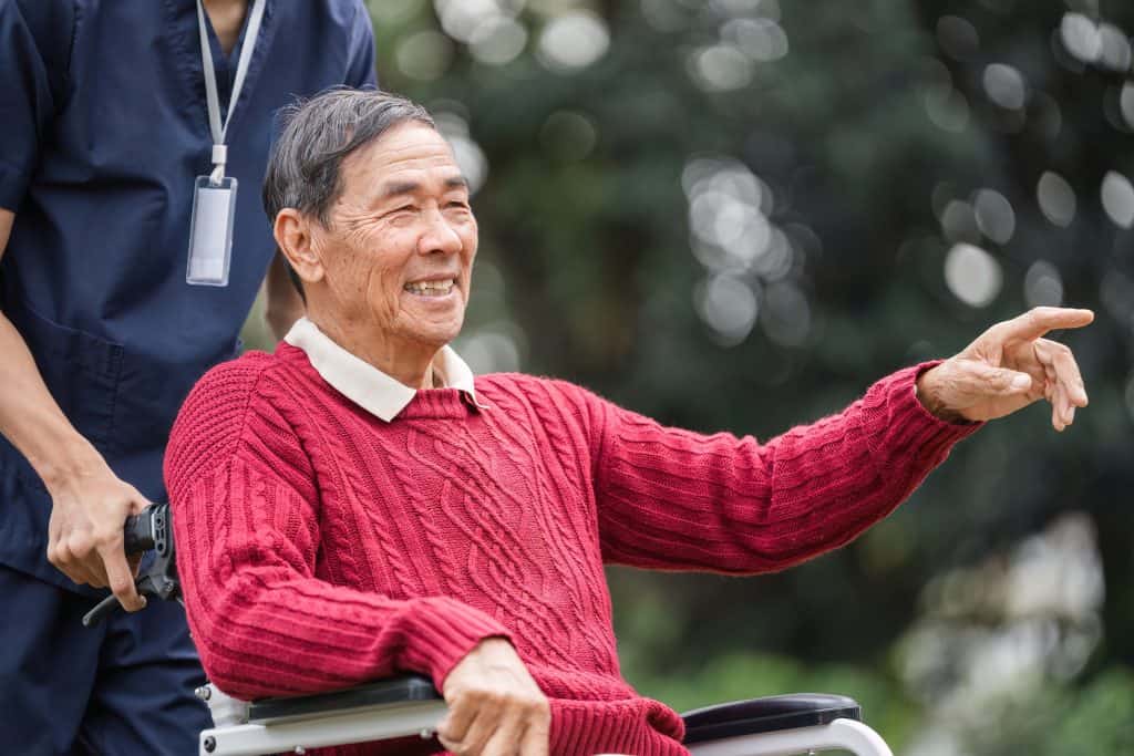 Elderly man with a warm smile happily gesturing while sitting in a wheelchair, receiving attentive care from his caregiver in a serene garden environment.